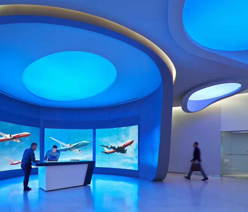 Two people use a touchscreen table in a blue-lit room with airplane screens; another walks by futuristic, curved walls.