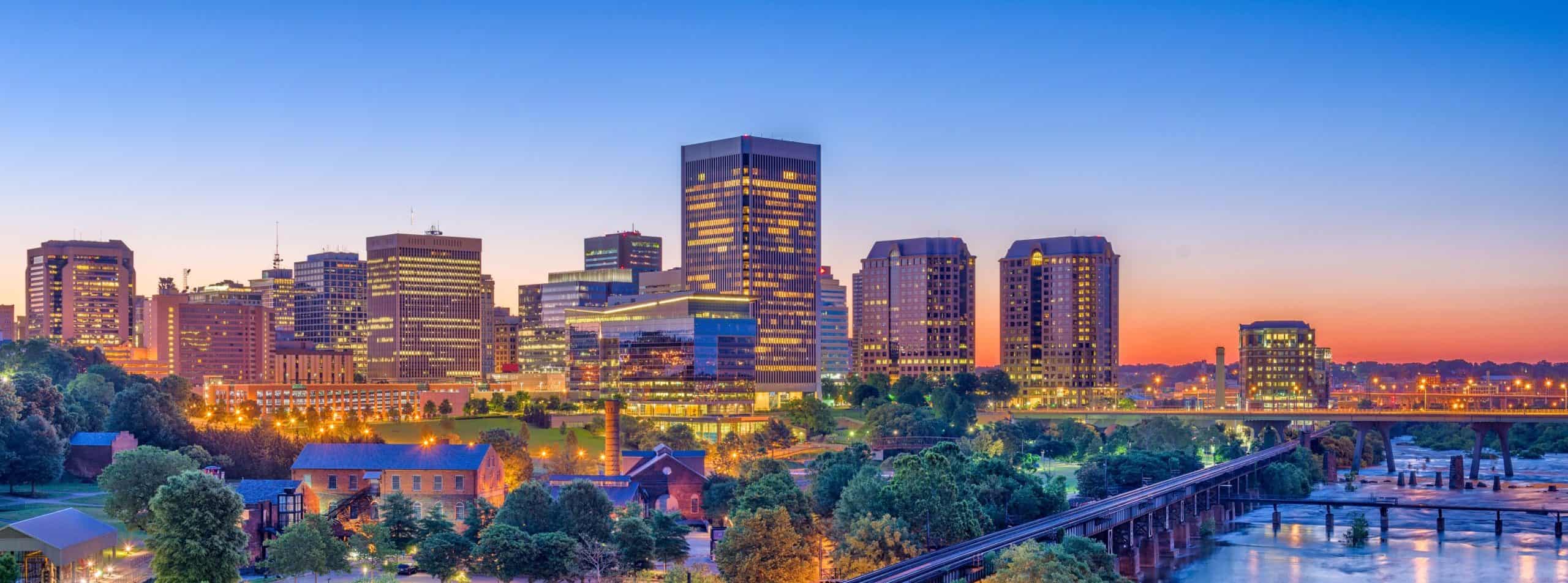 City skyline at dusk: illuminated skyscrapers, river & bridge, lush trees in foreground under an orange-to-blue sky.