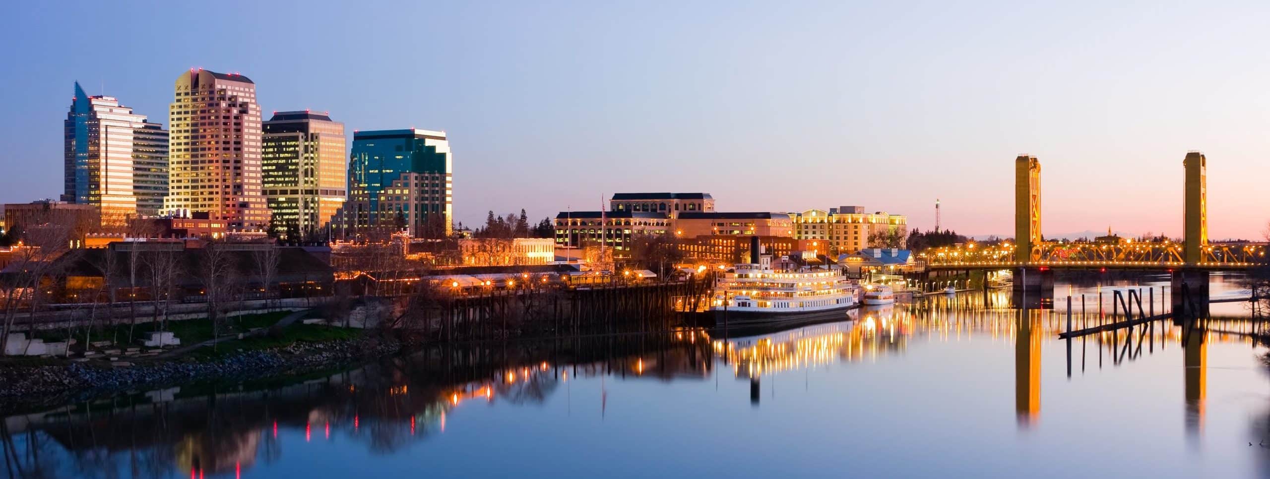 City skyline at dusk, illuminated buildings, river reflections, golden-lit bridge on the right.