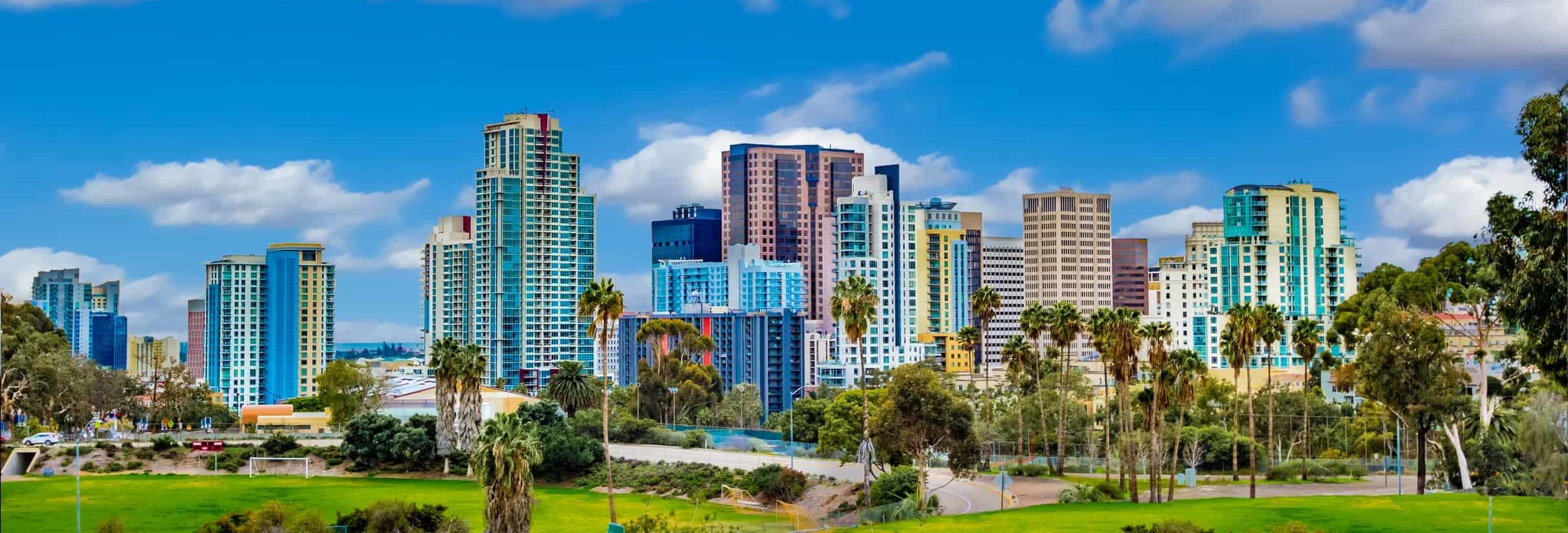 Modern city skyline with diverse high-rises under a blue sky, lush green park in foreground contrasts urban backdrop.