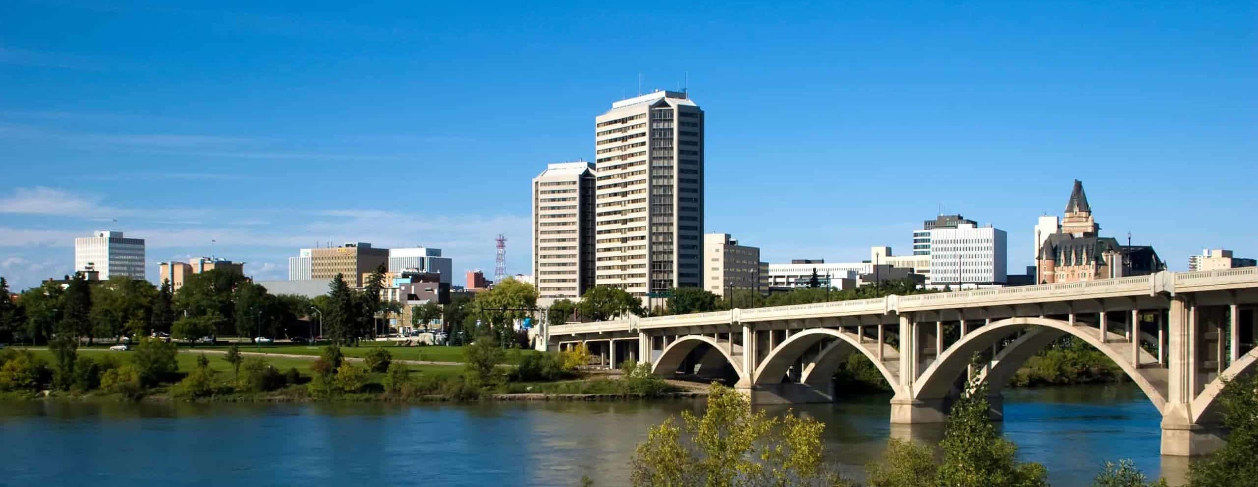 City skyline with modern high-rises, historic tower, blue sky; river and stone bridge in foreground, surrounded by trees.