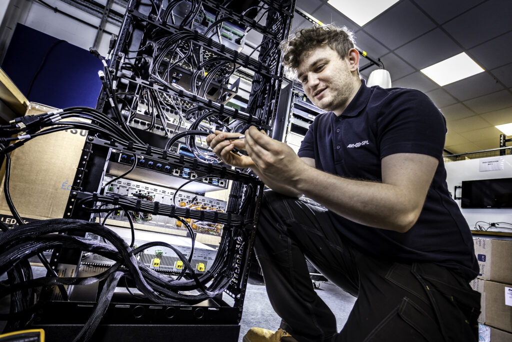 A technician in a black shirt examines cables by server racks in a tech-filled room with a tiled ceiling, creating an industrial vibe.