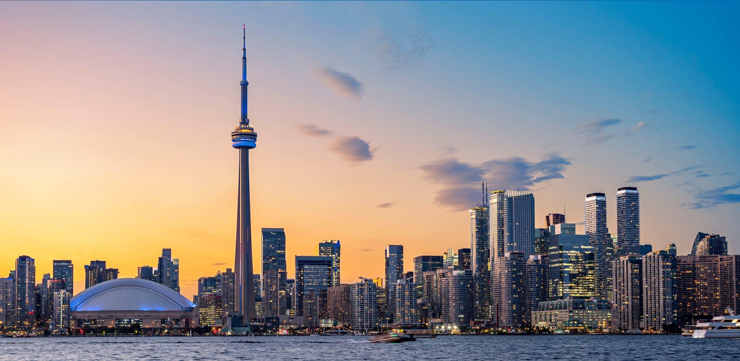 Toronto skyline at sunset: CN Tower, lit skyscrapers, colorful sky, and rippling Lake Ontario in the foreground.