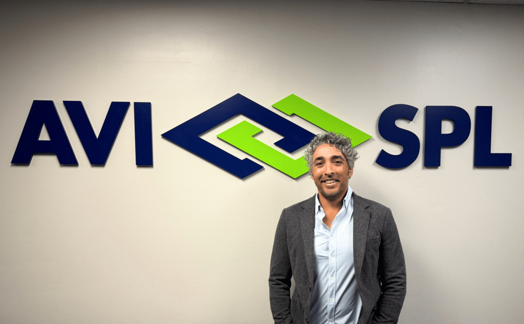 Gray-haired, bearded man in gray blazer and blue shirt smiles in front of AVI SPL logo with interlocking blue and green shapes.