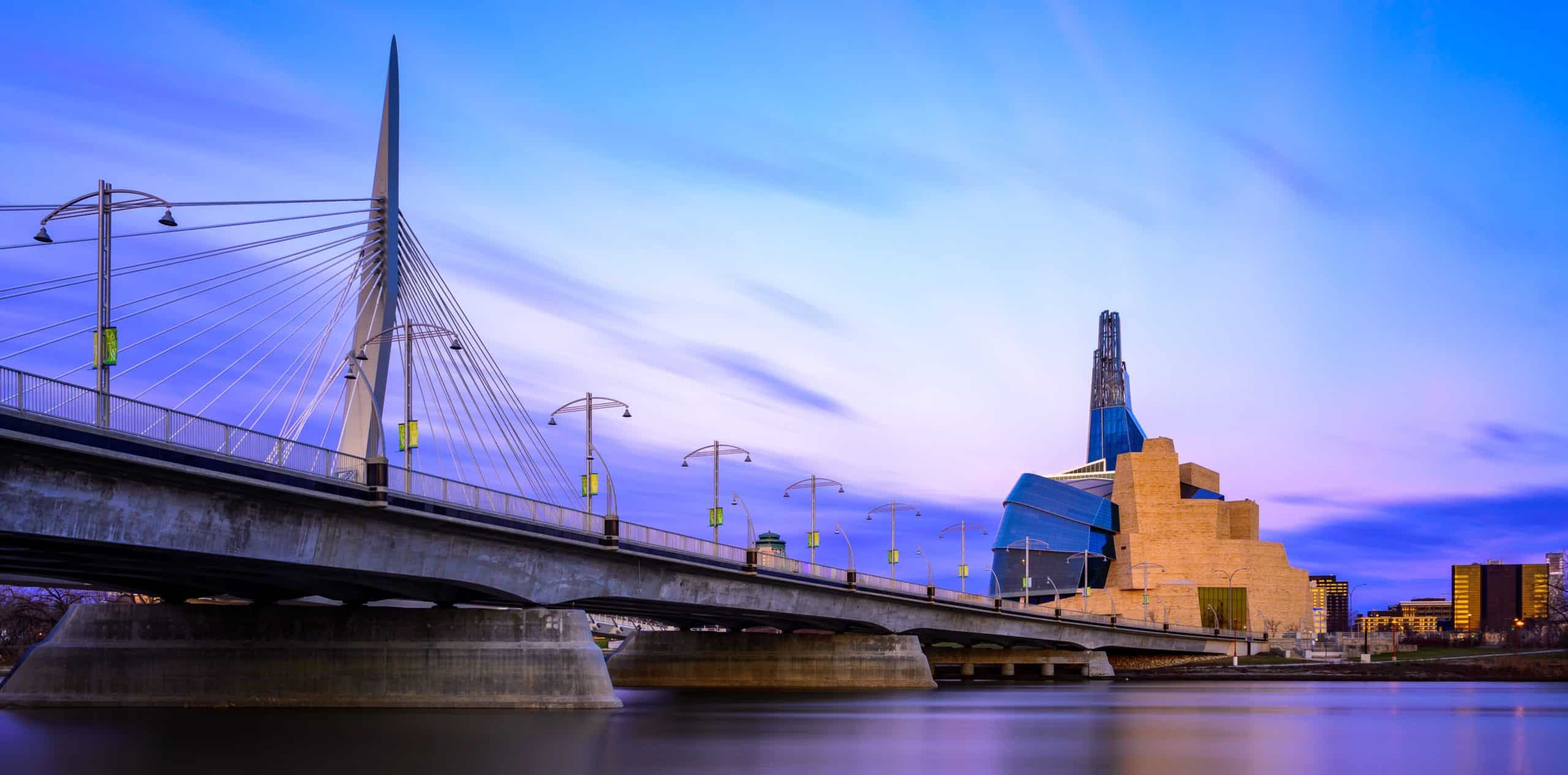 Esplanade Riel and Canadian Museum at dusk: bridge cables fan elegantly, museum aglow against serene evening sky.