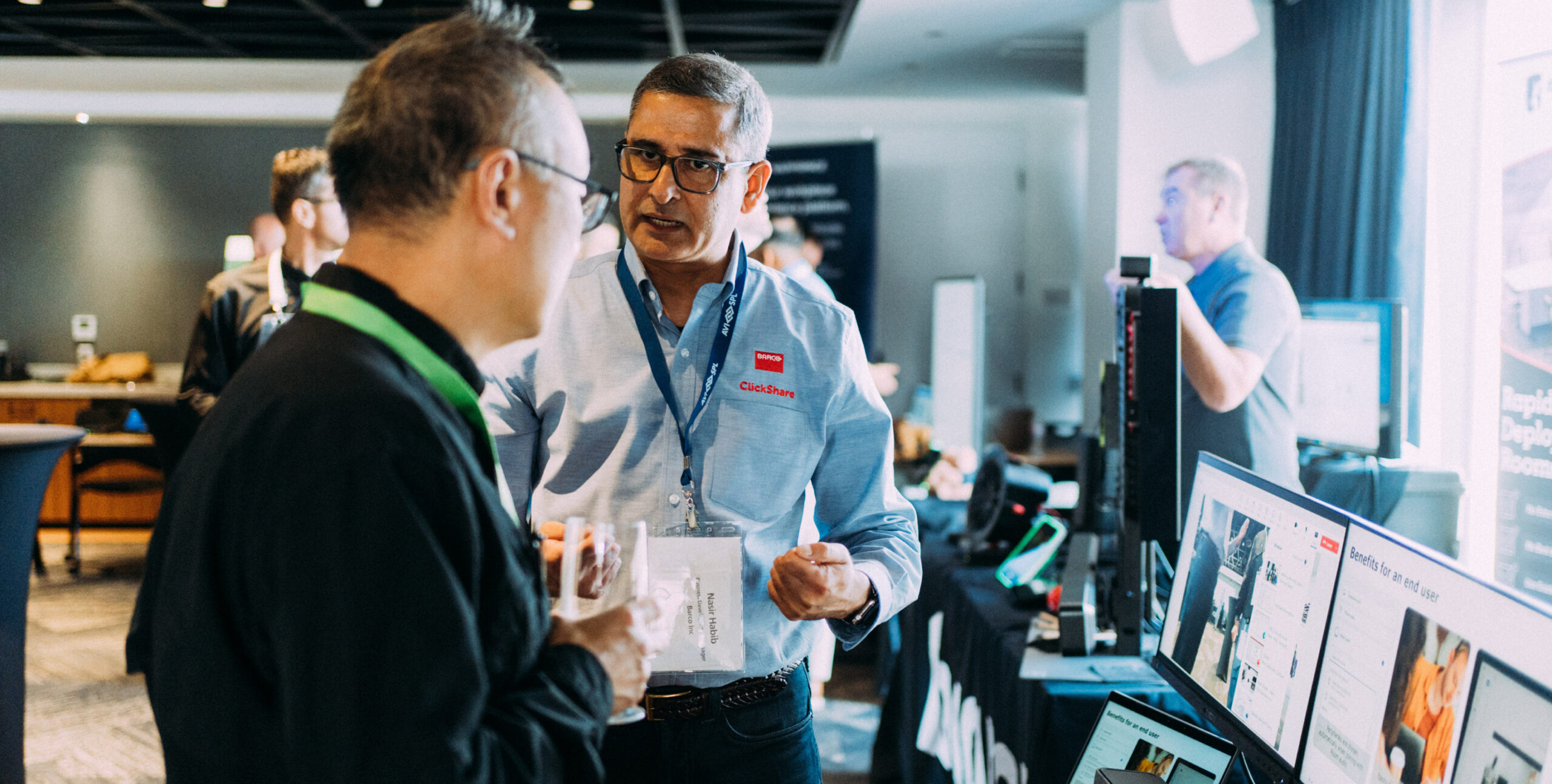 At a tech conference, two men chat; one with a badge and paper, the other with a drink. Screens and booths fill the background.