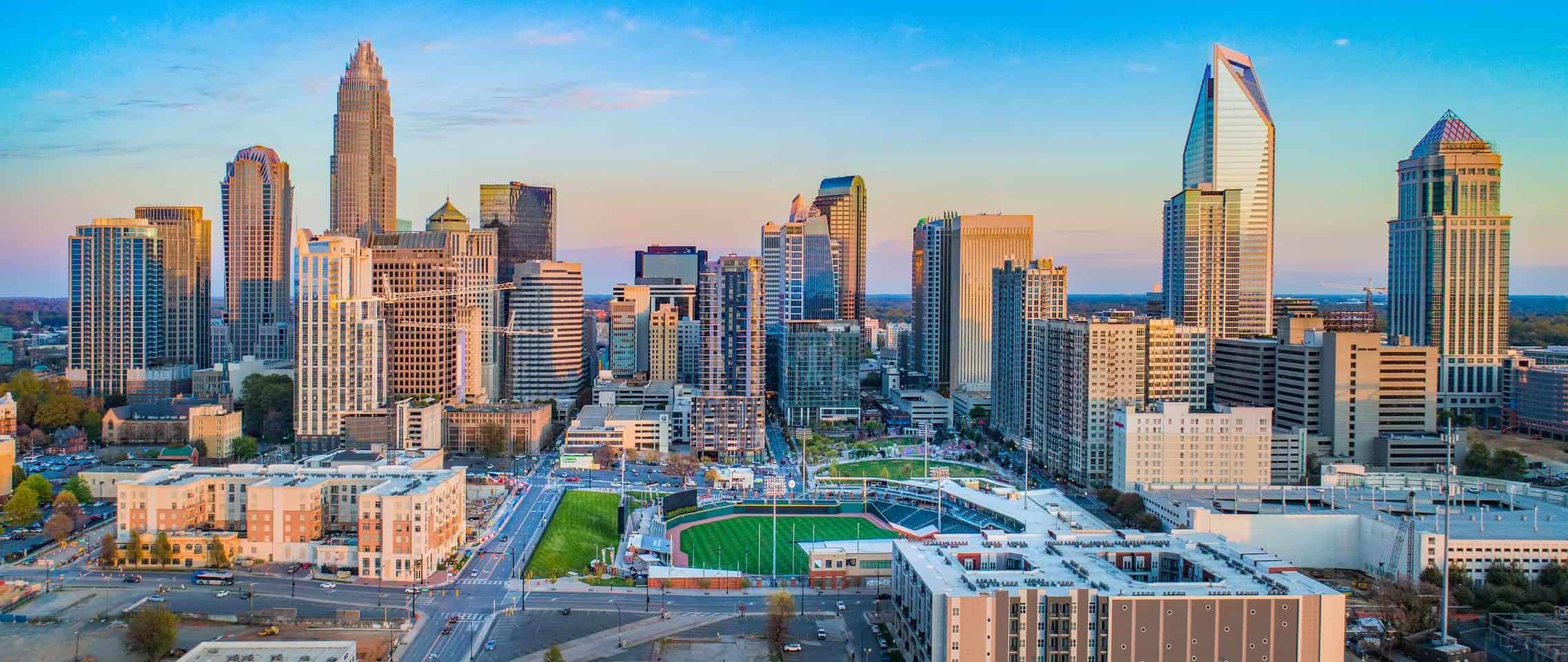 City skyline with skyscrapers under clear blue sky, surrounded by mixed-use buildings, a green sports field, and visible street patterns.