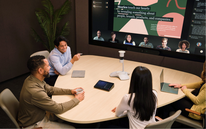 People at a conference table on a video call, laptops open. A screen shows virtual attendees and a presentation on storytelling.