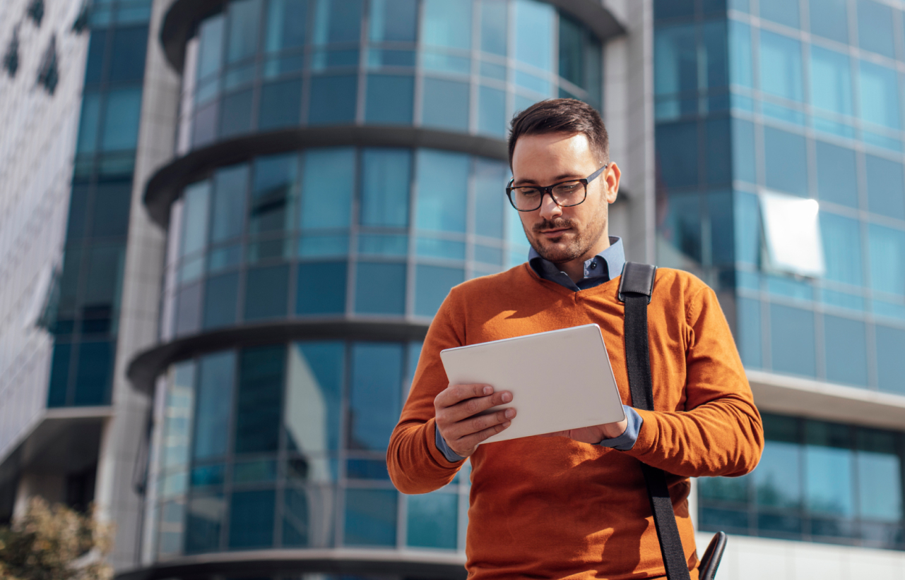 Man in orange sweater and glasses stands outside modern glass building, looking at tablet; shoulder bag slung across.