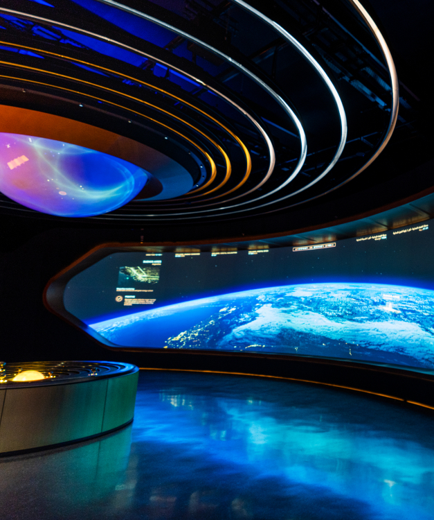 Futuristic room: curved screen shows Earth from space, ringed ceiling lights, control panel foreground; high-tech vibe.