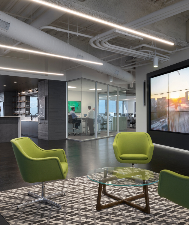 Modern office with green chairs, glass table on a rug; cityscape screen; people in a glass conference room; exposed ceiling ducts.