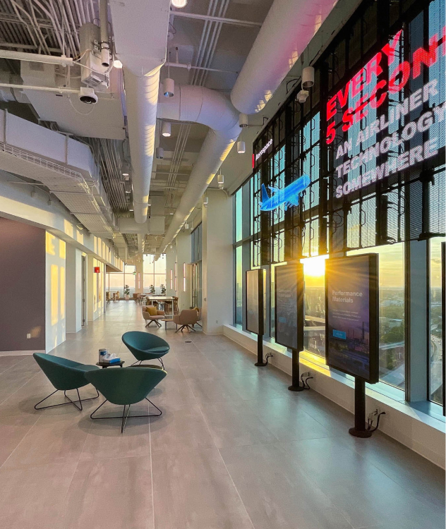 Modern office hallway with high ceilings, large windows, digital screens, stylish chairs, tables; sunlight highlights open design.