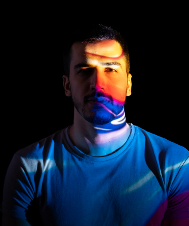 A person in a white shirt is lit by vibrant multicolored projections in a dark setting, emphasizing colors and shadows on them.