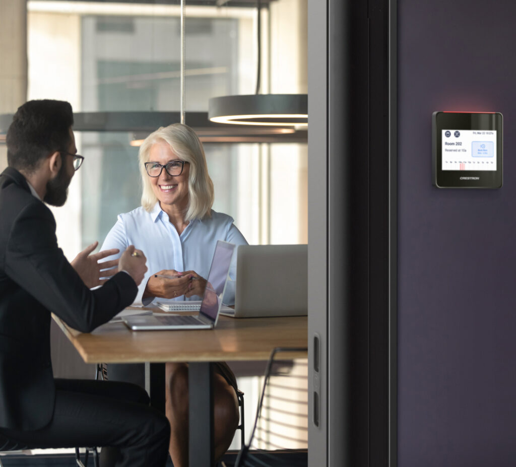 A man and woman chat at a table in a modern office, smiling with open laptops; meeting info shown on a nearby digital screen.