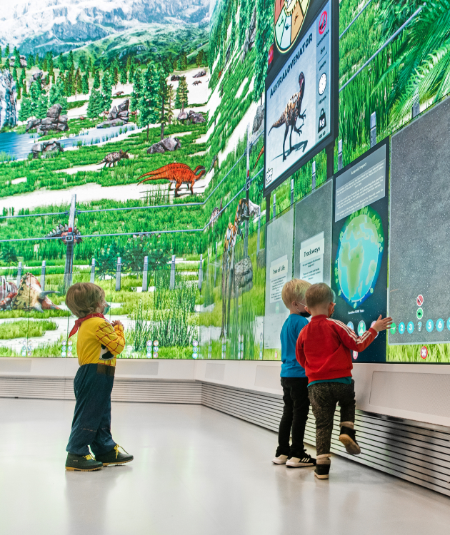 Kids interact with a digital screen showing a vivid prehistoric scene, with dinosaurs, greenery, and info panels. Theyre engaged and exploring.