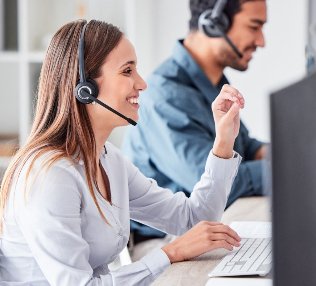 A woman smiles, gestures, and works at her computer in an office. A man behind her focuses on his monitor. Both wear headsets.