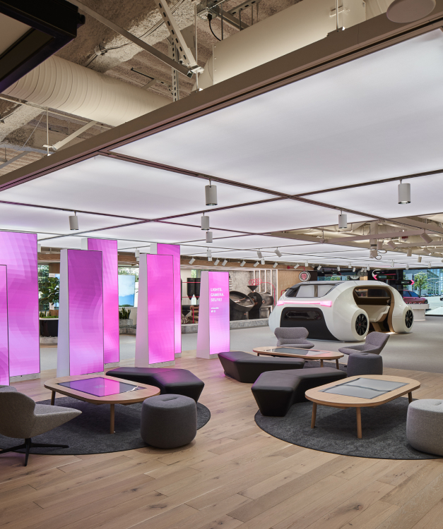 Modern office with purple-lit panels, circular seating, wood floors; futuristic round vehicle in background; exposed ducts and lights.