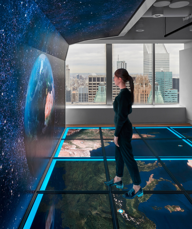 Person in room with Earth on screen, neon blue floor maps, cityscape visible through windows.