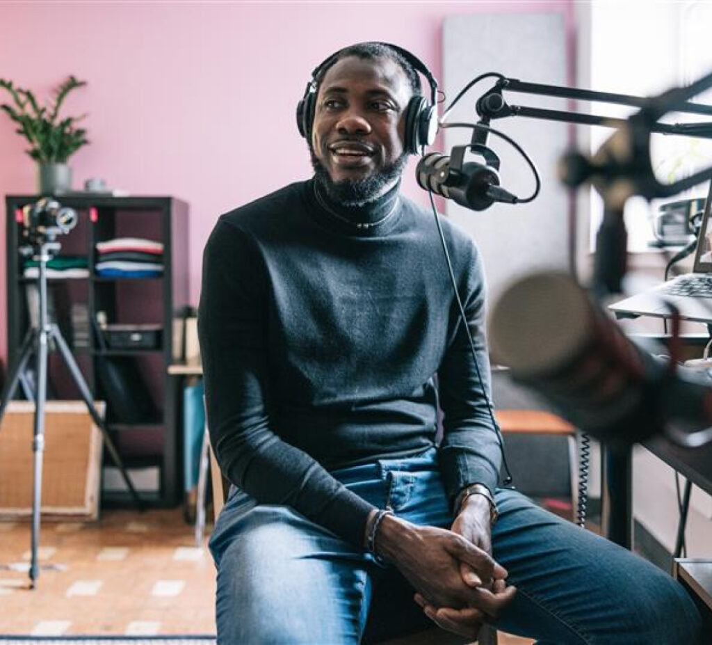 Man in headphones at studio mic, black sweater/jeans, pink wall, plant & shelves in background.