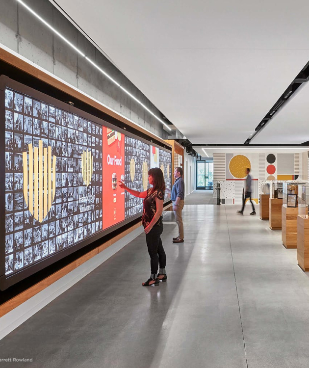 People engage with a digital display wall in a modern space, while others explore exhibits in the background.