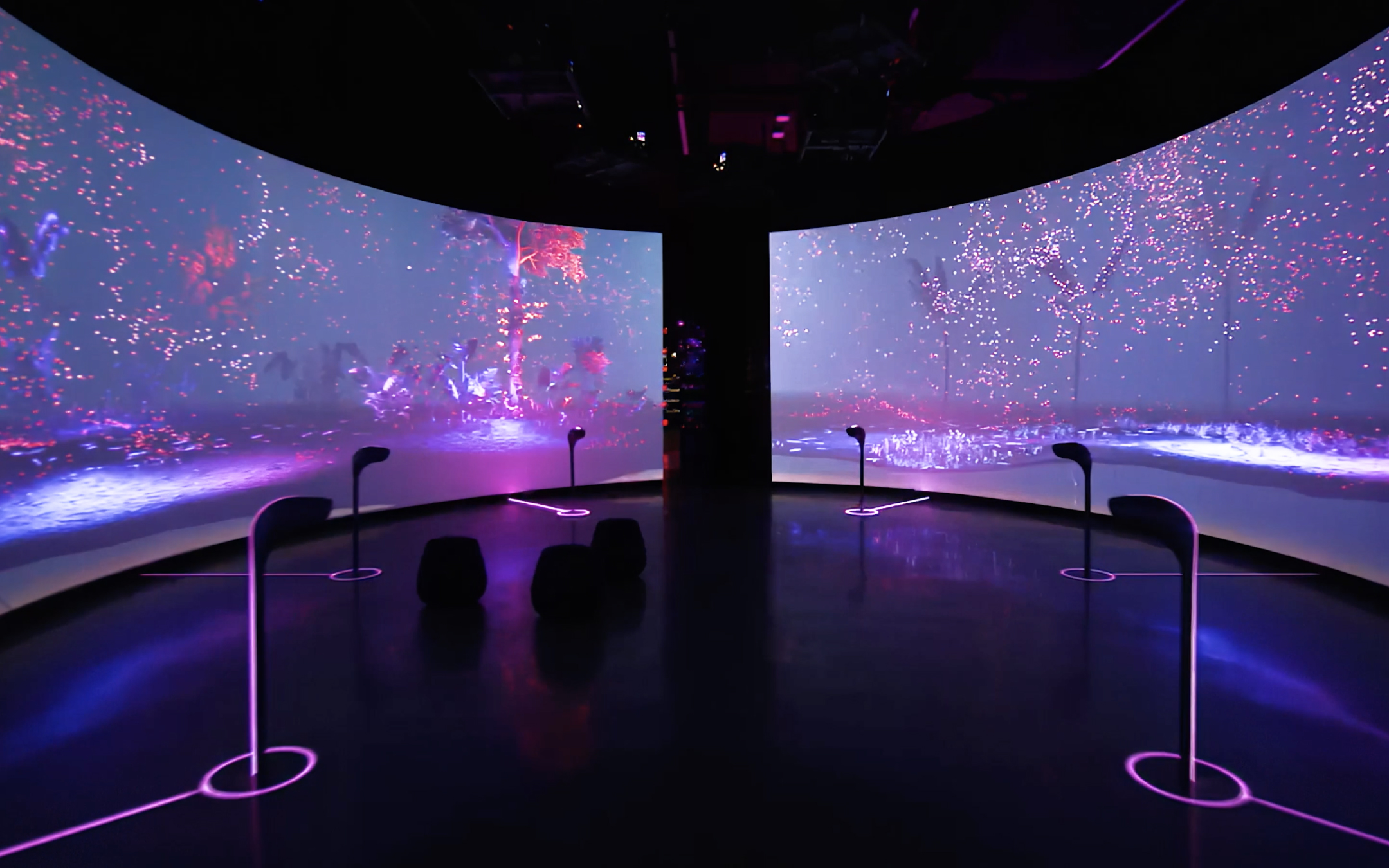 A dark room with curved screens shows vibrant pink/purple art. Black stools and speakers create an immersive, futuristic vibe.