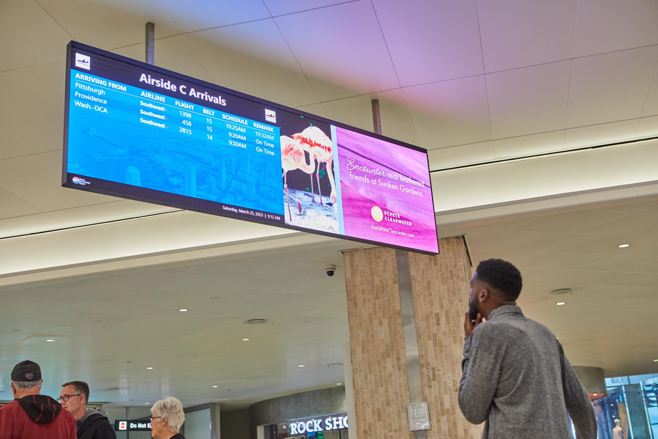 Man views arrivals board; ad for Grenadas beaches & culture nearby. Travelers in background.