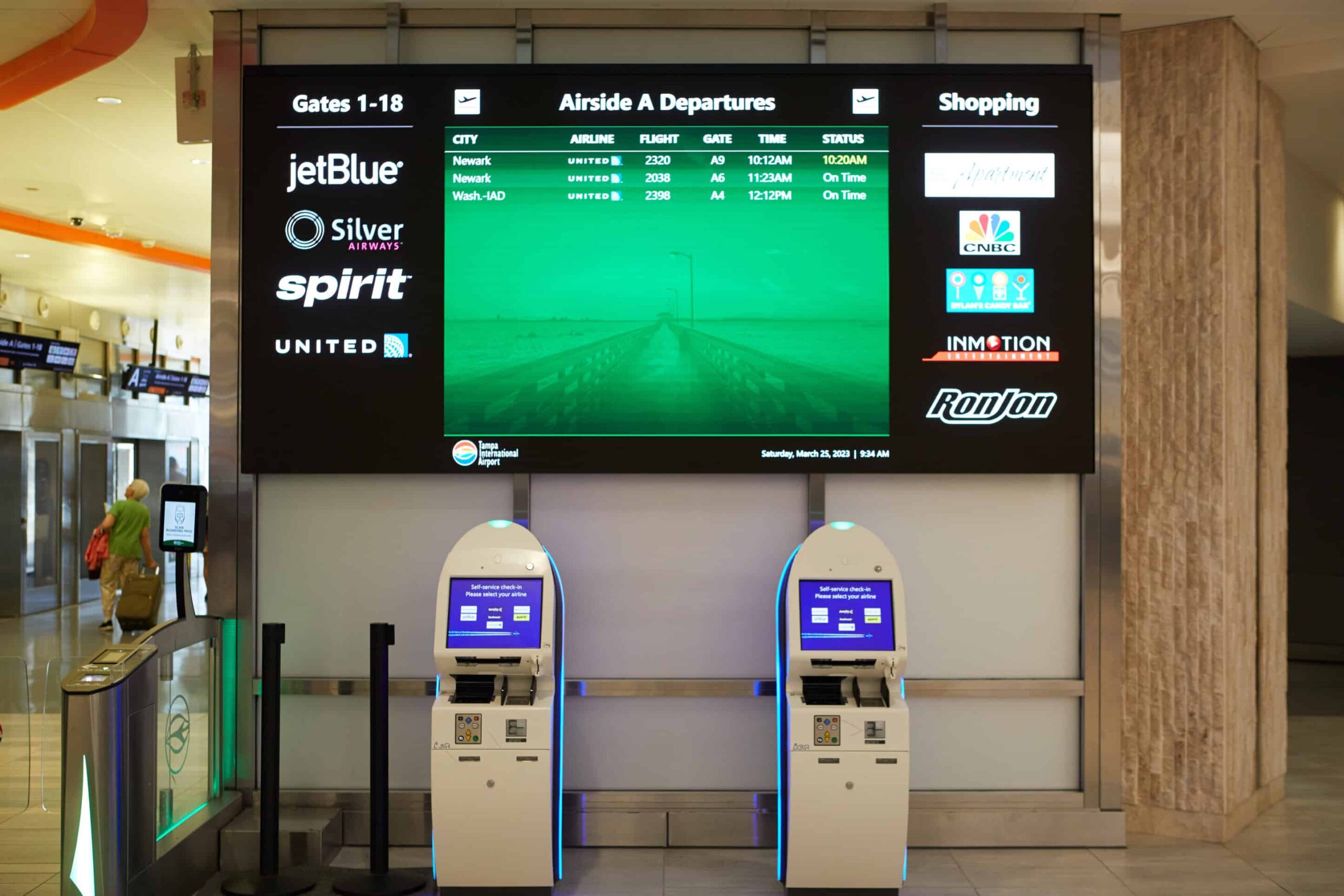 Airport terminal with digital departure board, JetBlue & Spirit info, self-serve kiosks, shopping & food logos visible.