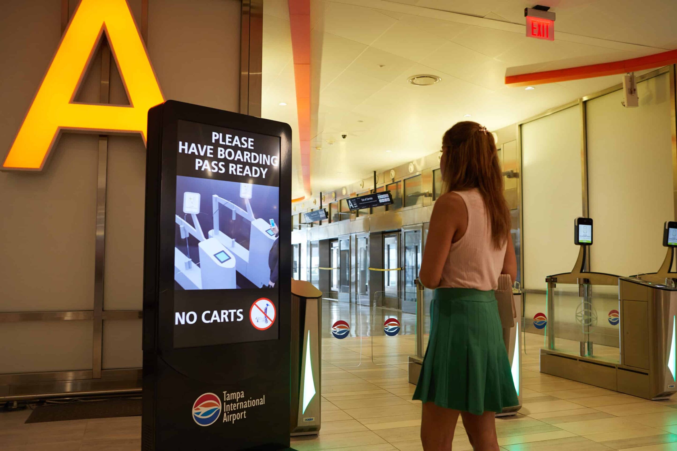 A woman stands before a digital sign at Tampa Airport, instructed to have a boarding pass ready; carts prohibited.