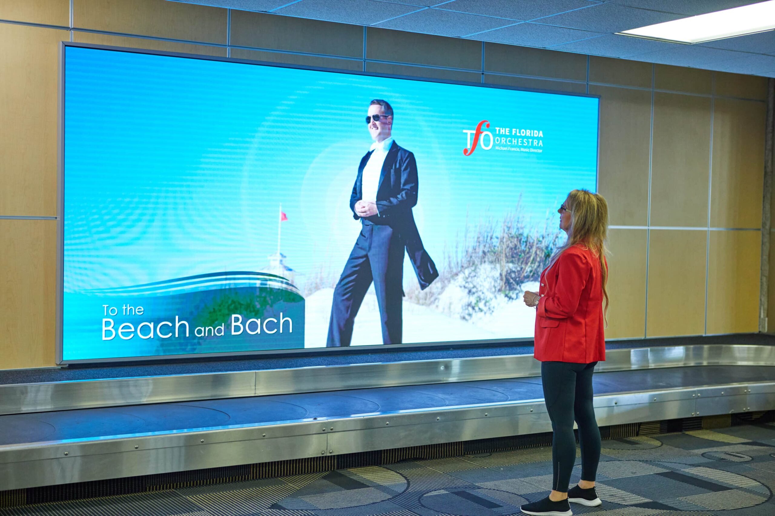 A woman in a red jacket stands before a large airport ad displaying The Florida Orchestra and To the Beach and Bach.