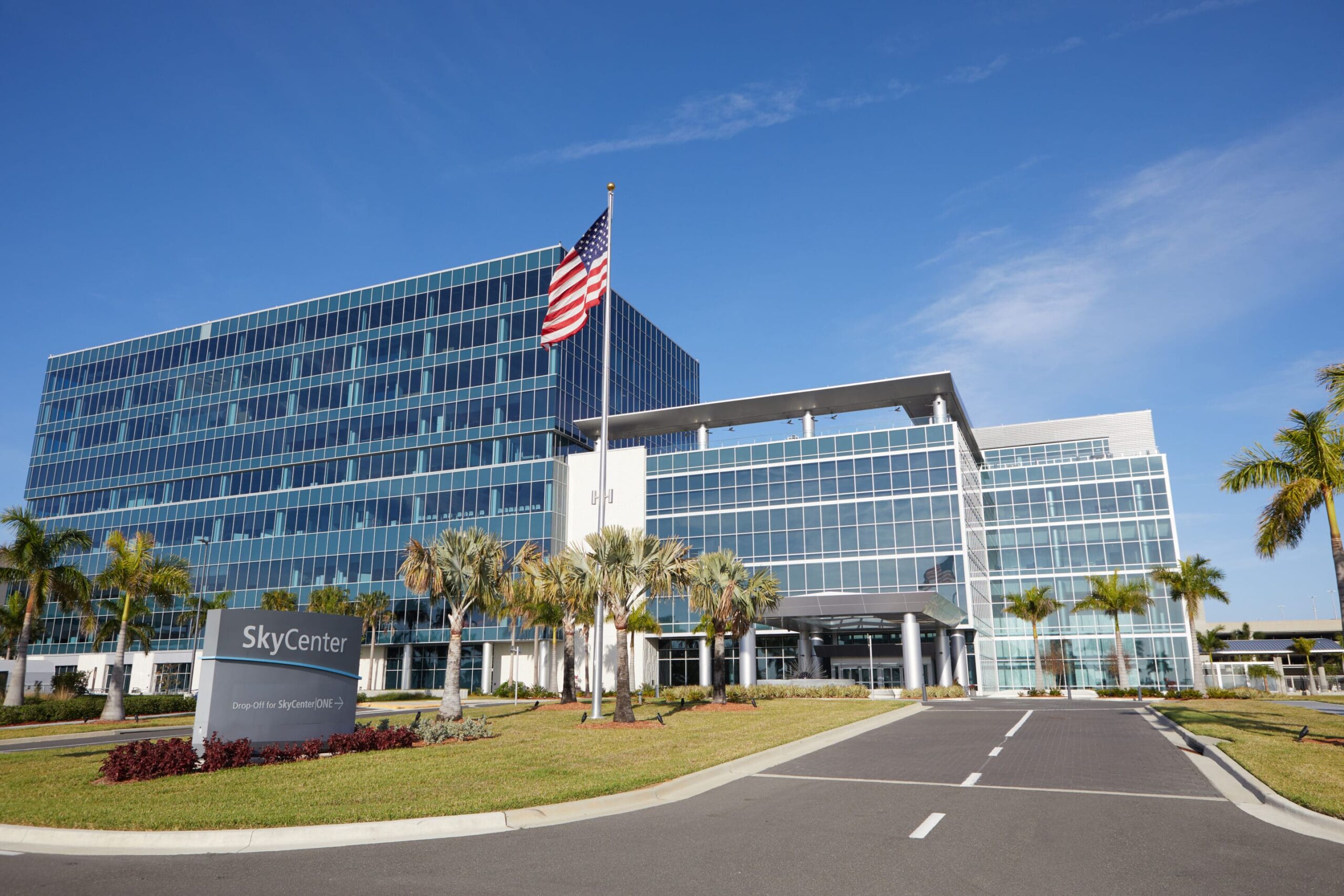 SkyCenter sign on a modern glass office, American flag nearby, palm trees, manicured lawn, clear blue sky, road to entrance.