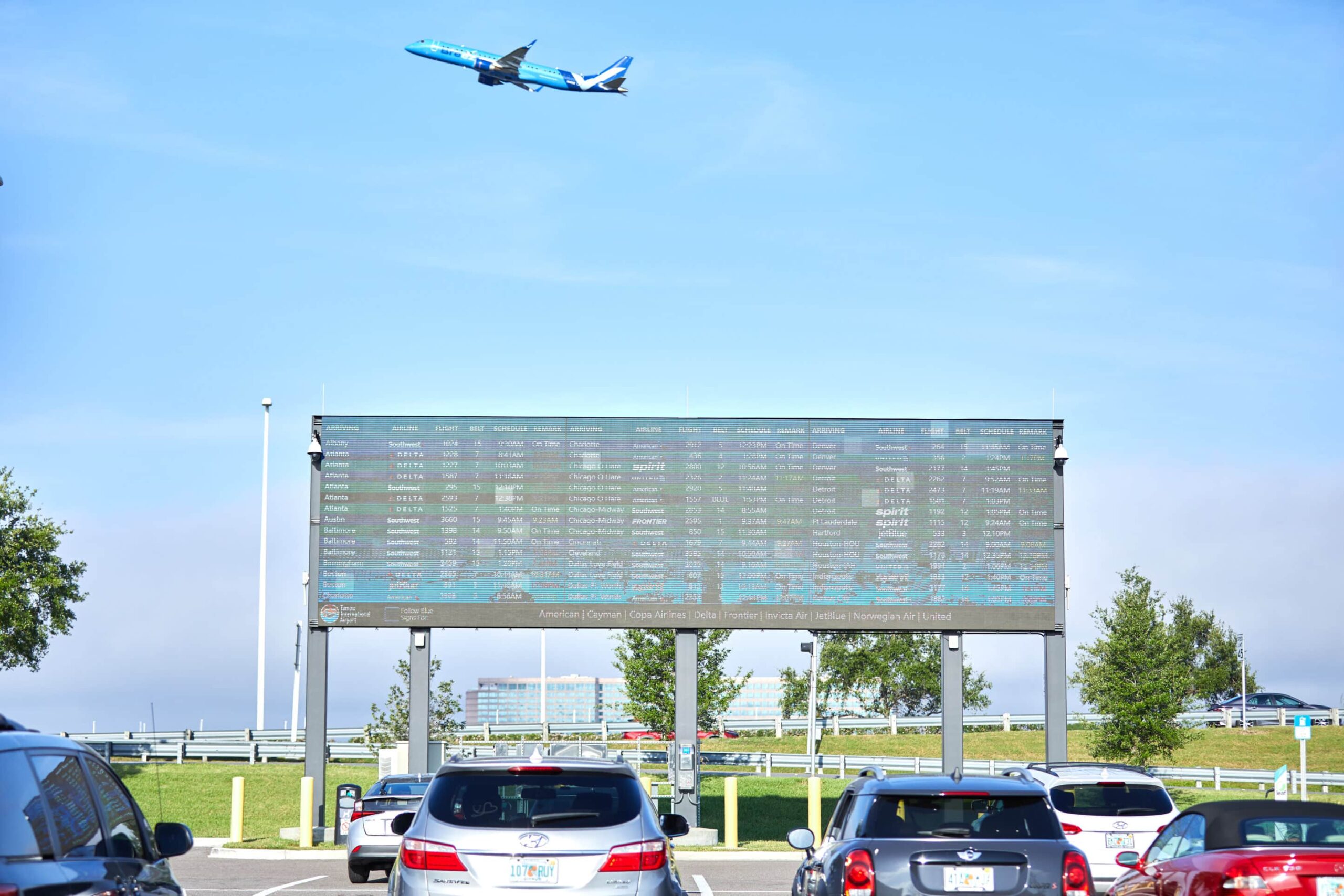 Cars in a lot with a display board showing flights, plane in clear sky above, trees and a building visible.