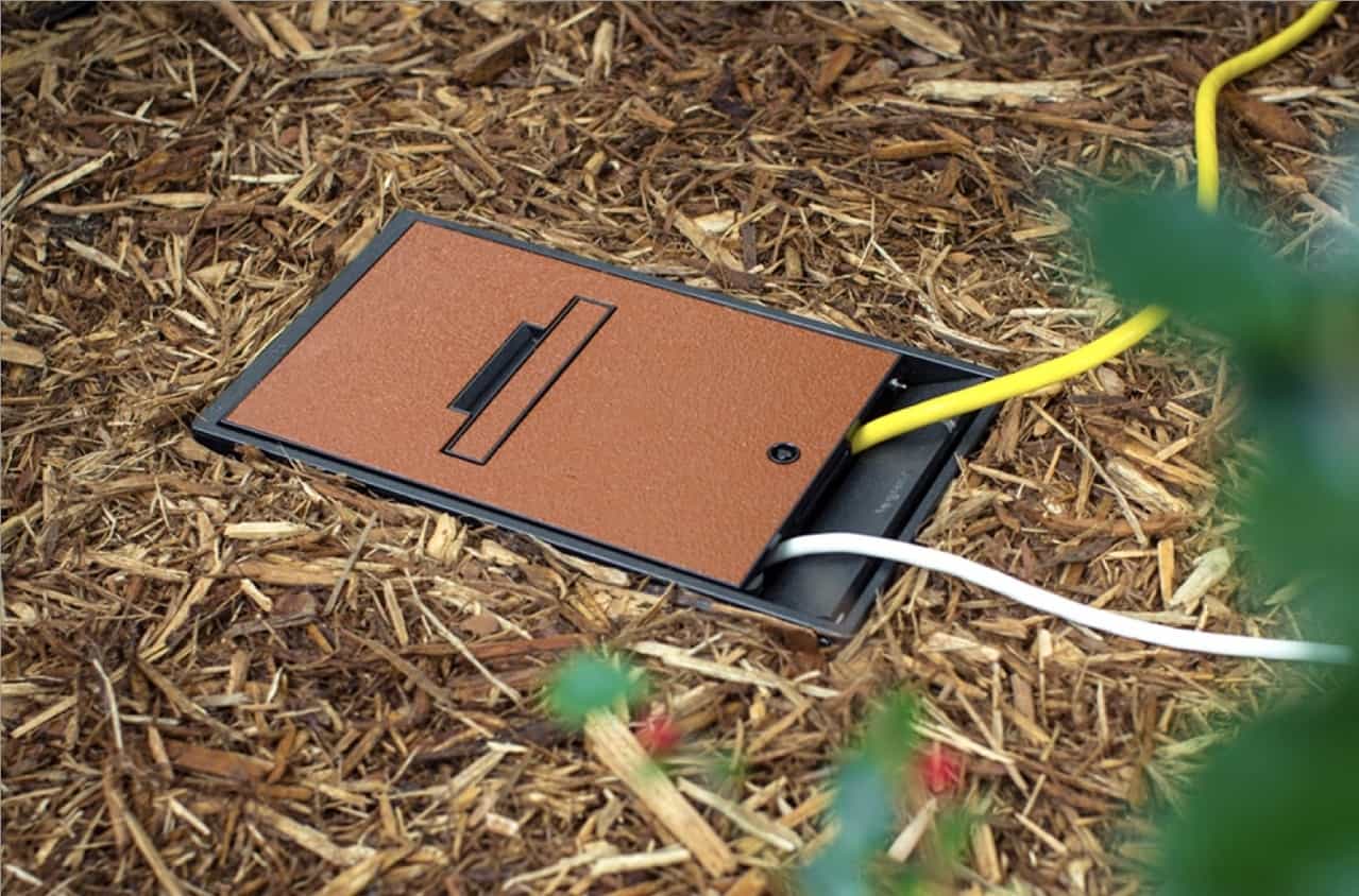 A brown power outlet cover in mulch with yellow/white cables; green plant partly visible on the right.