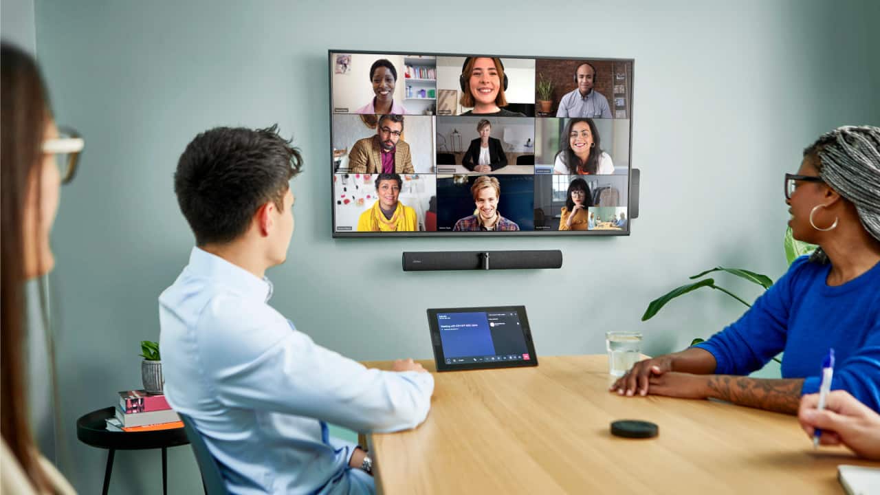 Four people sit around a table for a video call, displayed in grid view. A remote and tablet are also on the table.