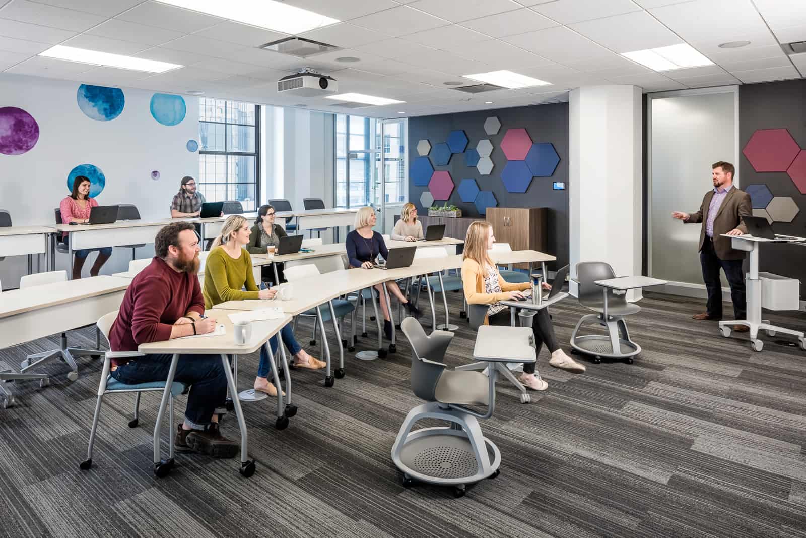 A modern classroom with students at desks in rows, using laptops, listening to an instructor. Hexagonal wall art and large windows.