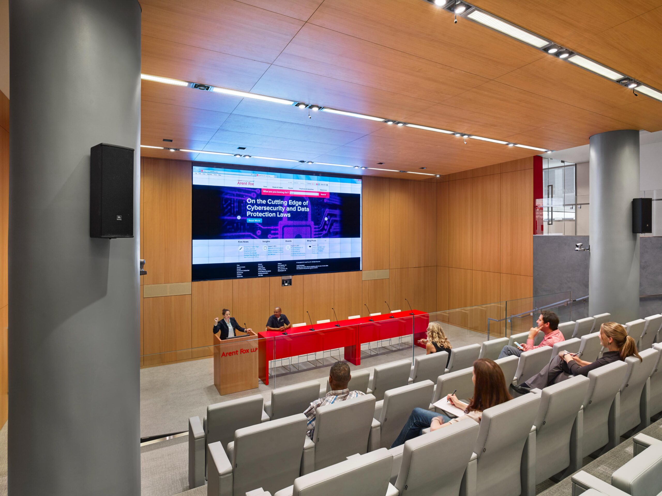 Modern lecture hall with digital screen, attendees on gray chairs taking notes. Speaker/assistant at red table, wood panel walls.