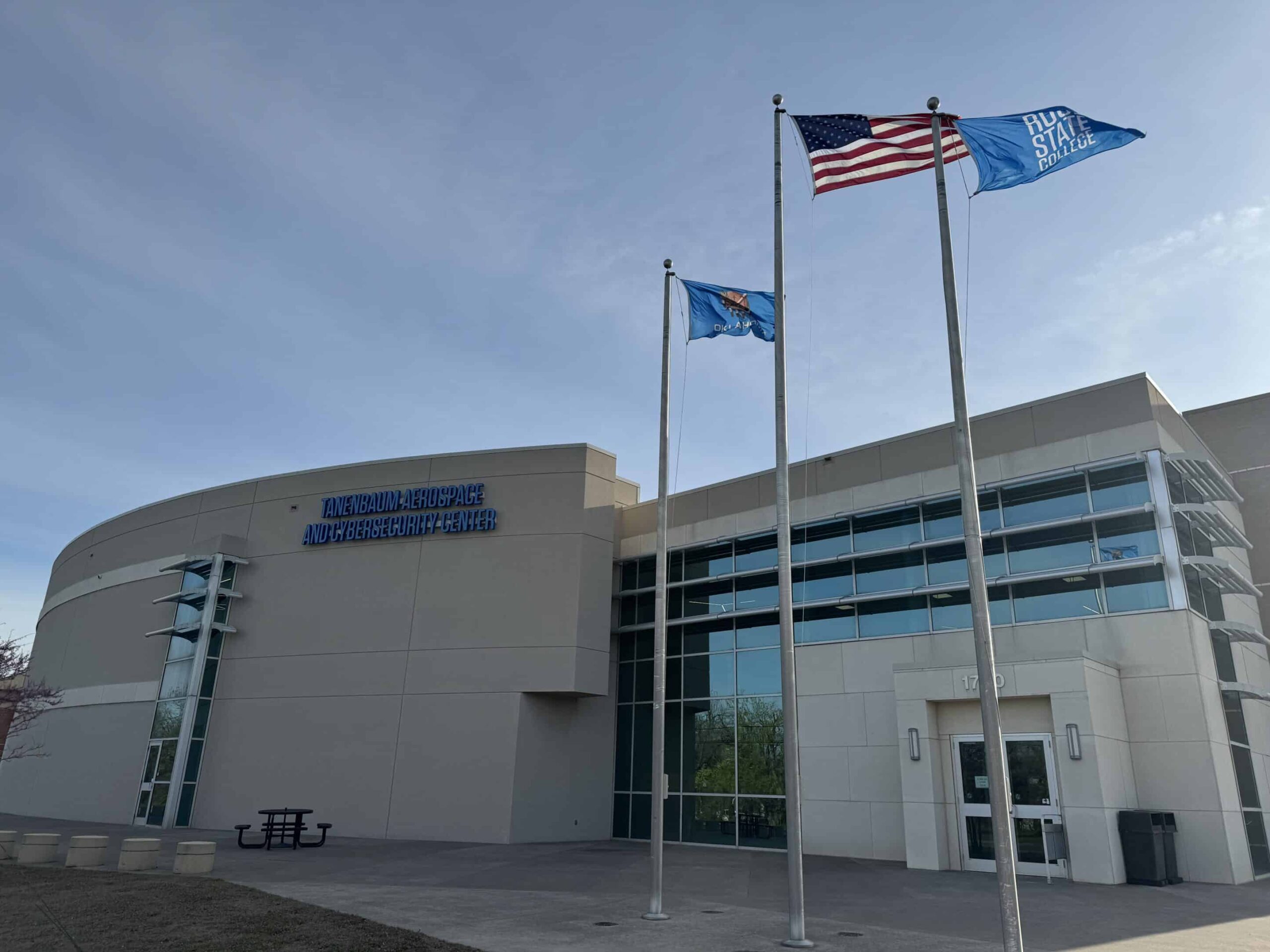 Modern building with flags: American, blue state, and blue with white text; large windows, curved facade, stairs, bench visible.