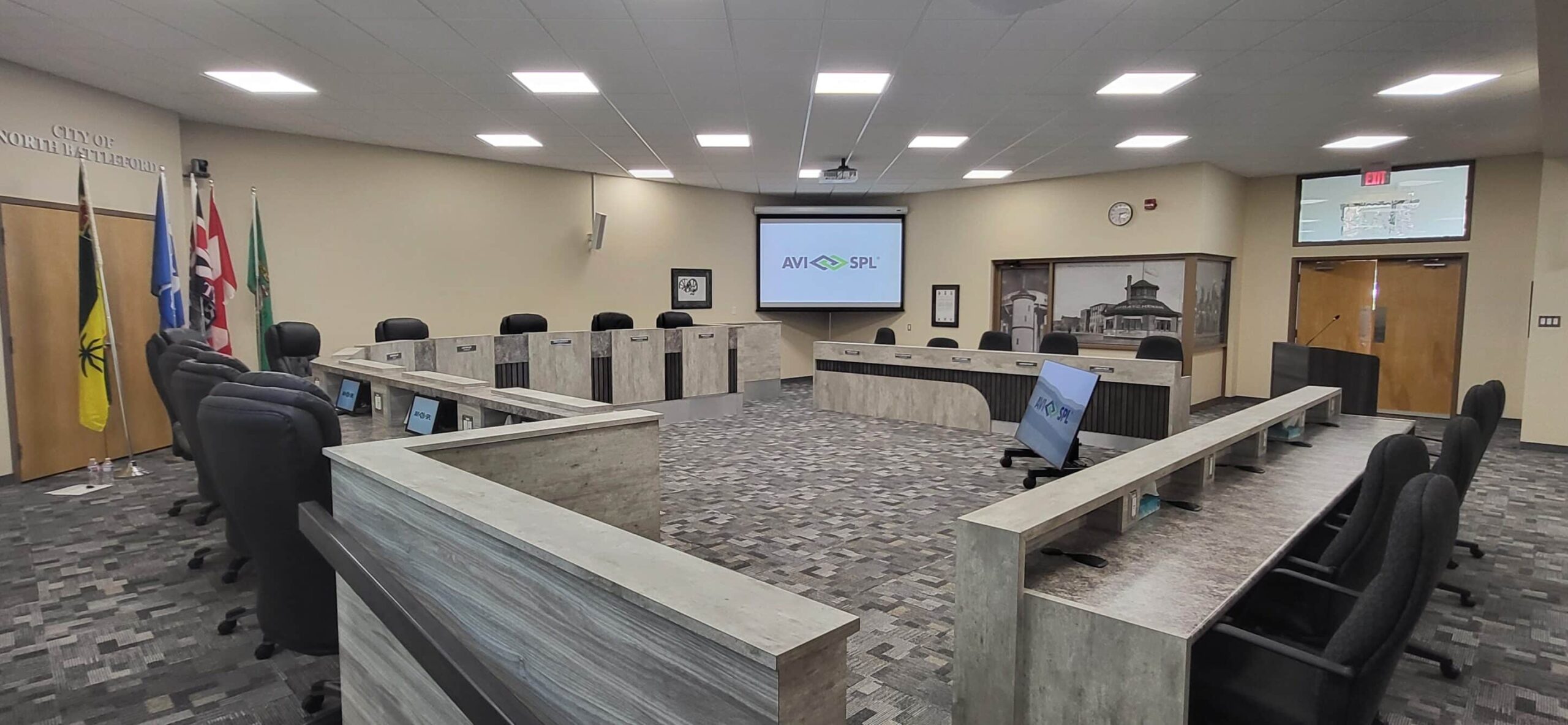 Modern council chamber: U-shaped desks, chairs, flags in corner, AVI-SPL on screen; well-lit, neutral tones, carpeted floor.