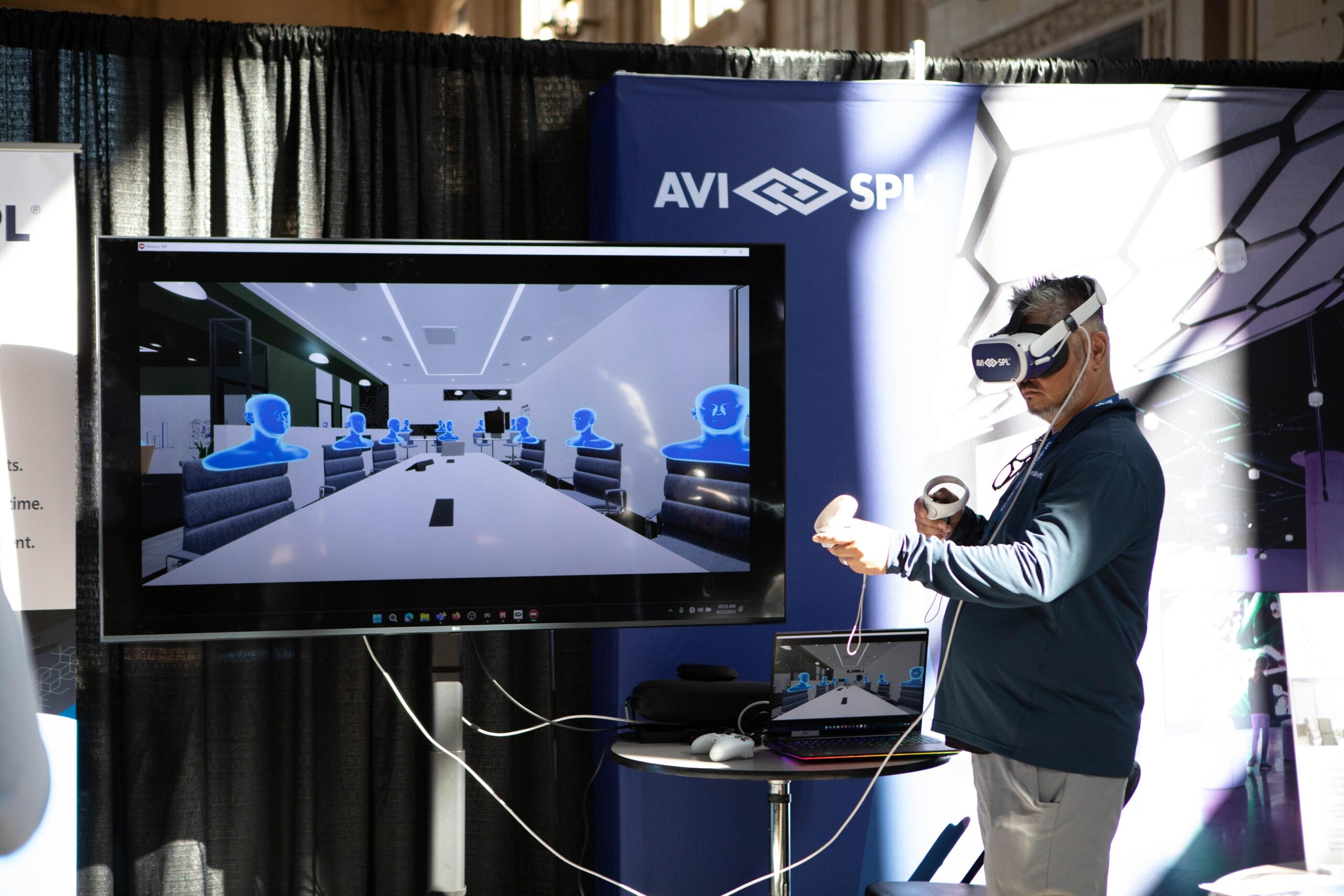 A person with a VR headset and controllers joins a virtual meeting at a tech exhibit booth with banners and a connected laptop.