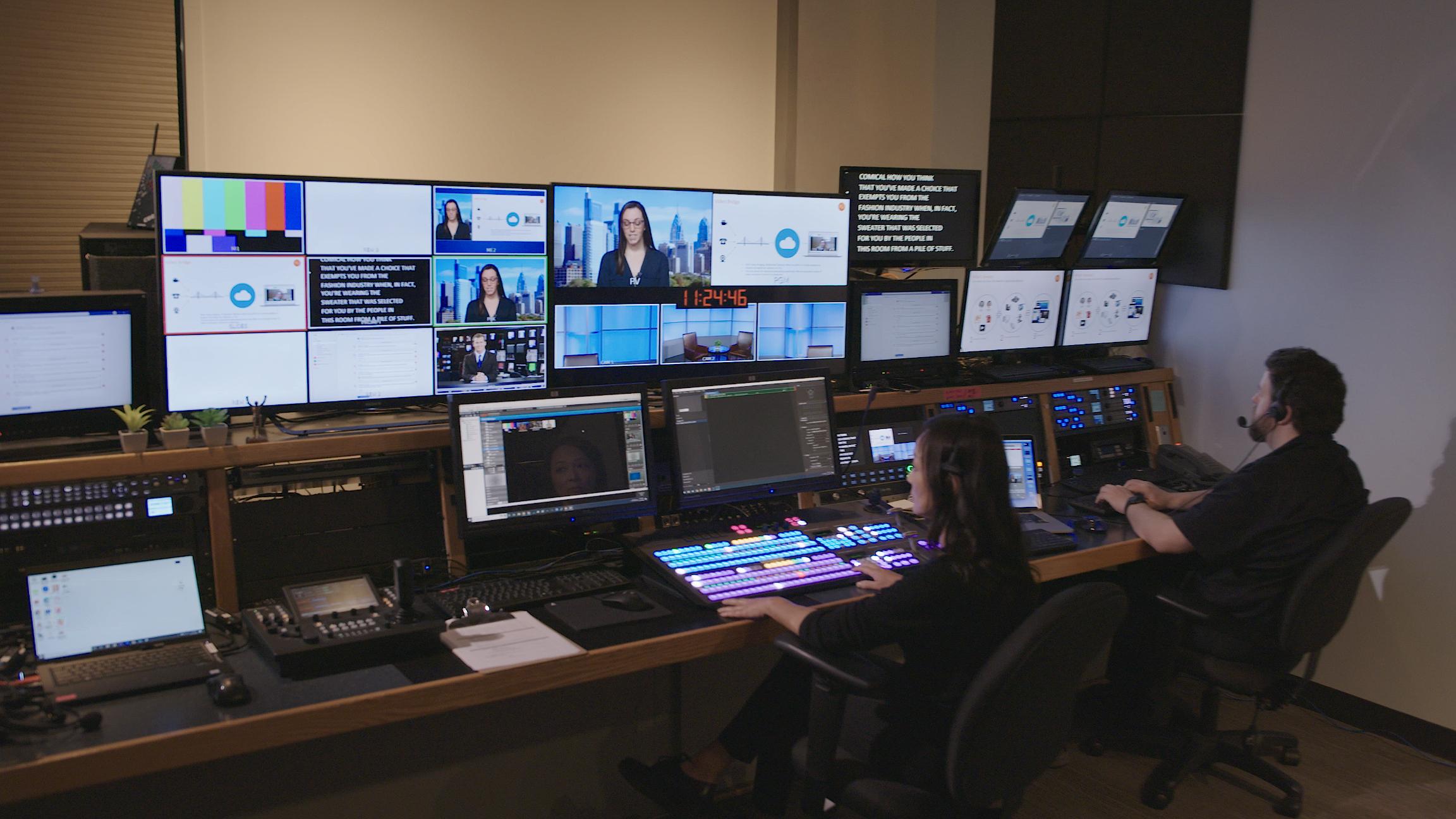 Two operators at a control room desk, surrounded by screens showing video feeds. Busy, high-tech atmosphere with studio lights visible.