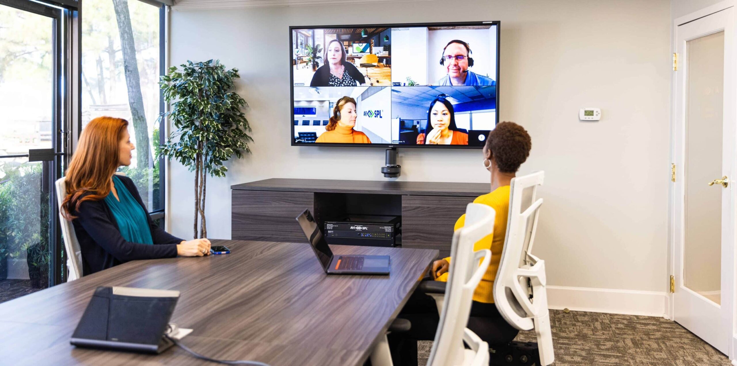 Two people at a conference table on a video call with five others displayed on screen; a laptop and notepad on the table.