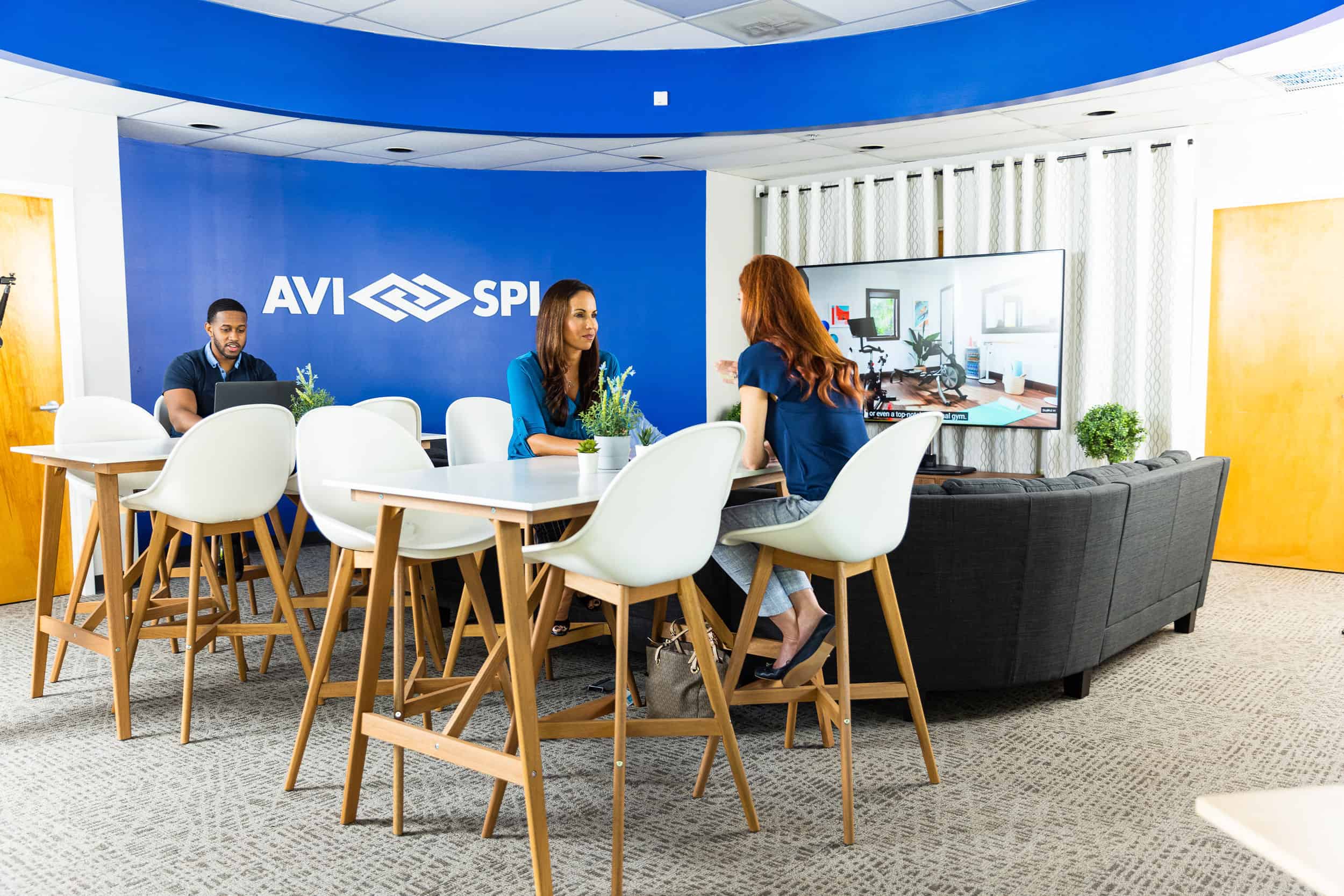 Three people in a modern office lounge with a video conference on TV; blue and white decor and logo on the wall.