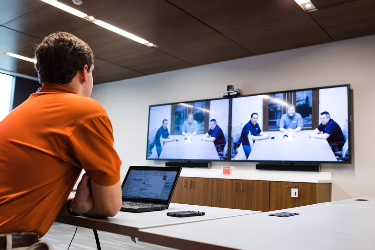 An orange-shirted person at a table watches a video call featuring four people in a modern, professional setting.