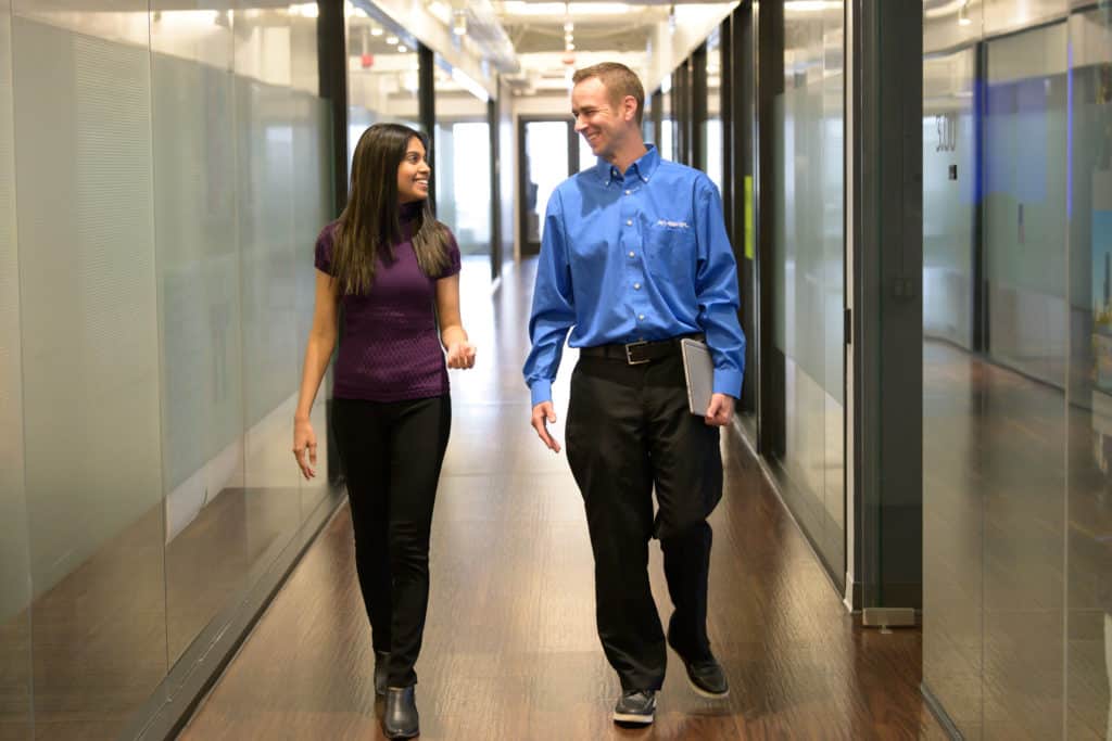 Two people walk and talk in a bright hallway, woman in purple shirt/black pants, man in blue shirt/black pants, engaged happily.