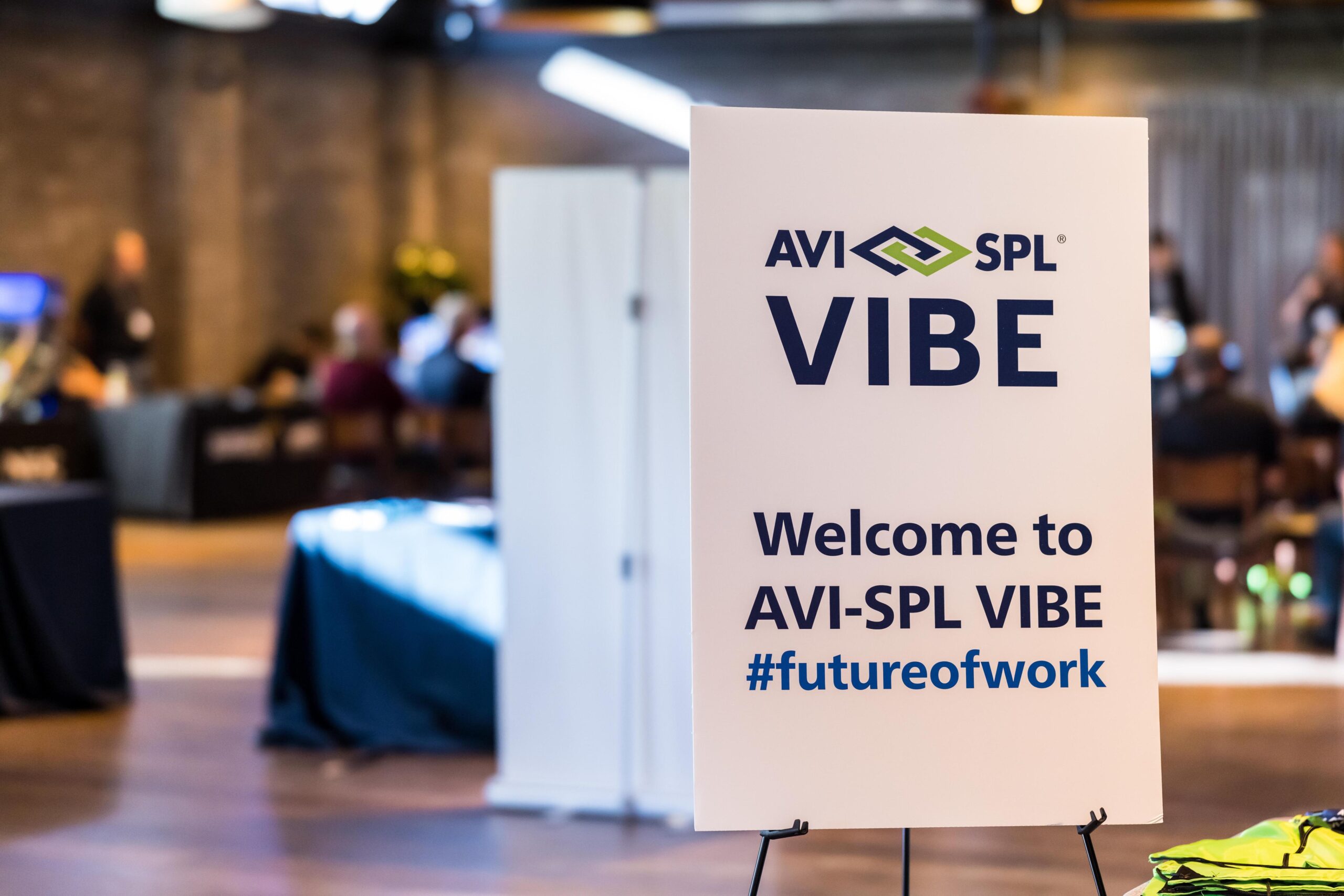 AVI-SPL VIBE sign: Welcome to AVI-SPL VIBE #futureofwork. Bright, modern event space with seated attendees.