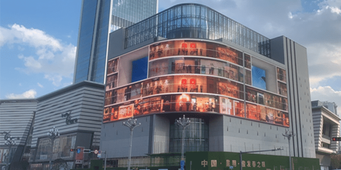 Modern multi-story building with digital displays, glass/steel elements; high-rise tower in background under partly cloudy sky.