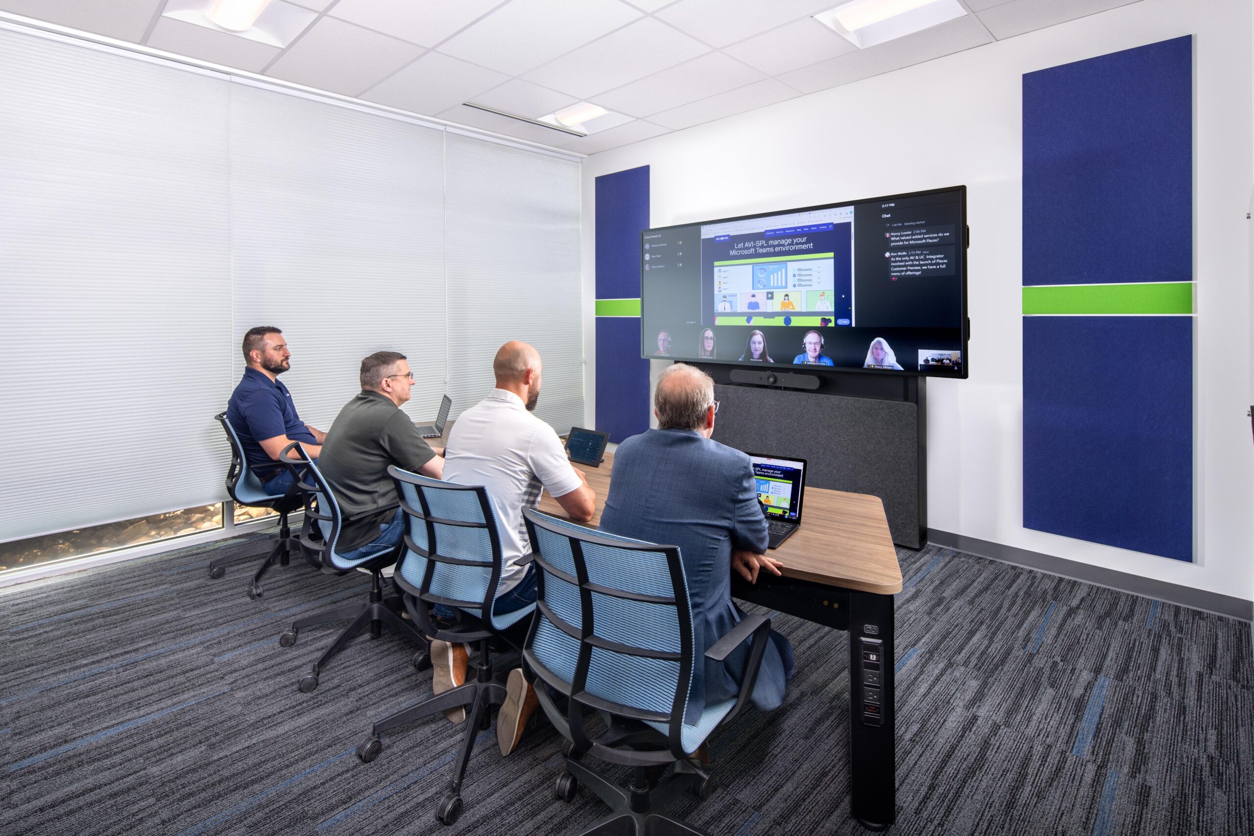 Four people sit at a conference table facing a large screen for a video call in a sleek, well-lit room with blue accents.