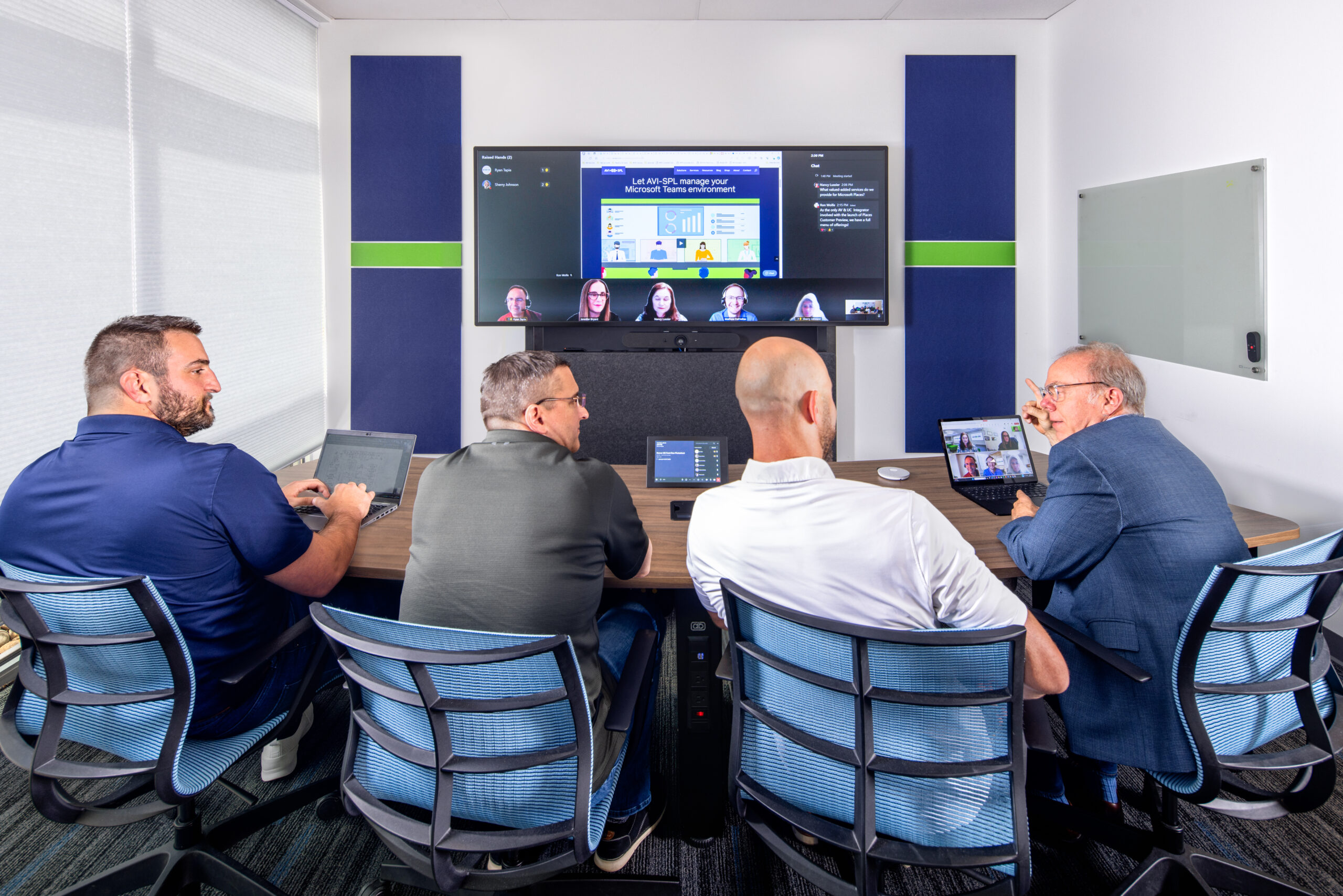 Five people at a conference table on a video call; large screen shows remote participants, room is bright and modern.