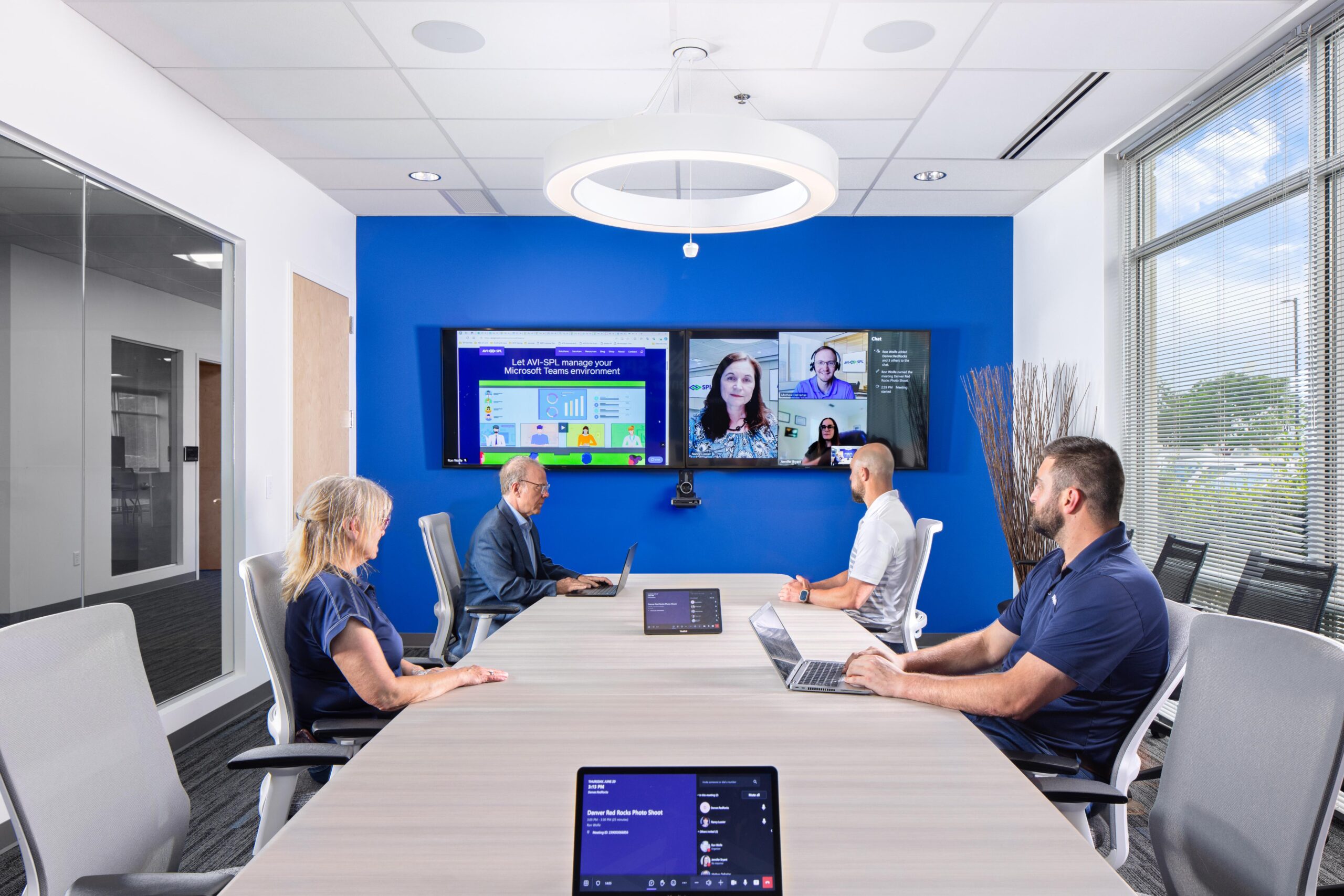 Five people meet virtually in a modern room: three at a table with laptops, two on screen. Tablets lie around; large window on right.