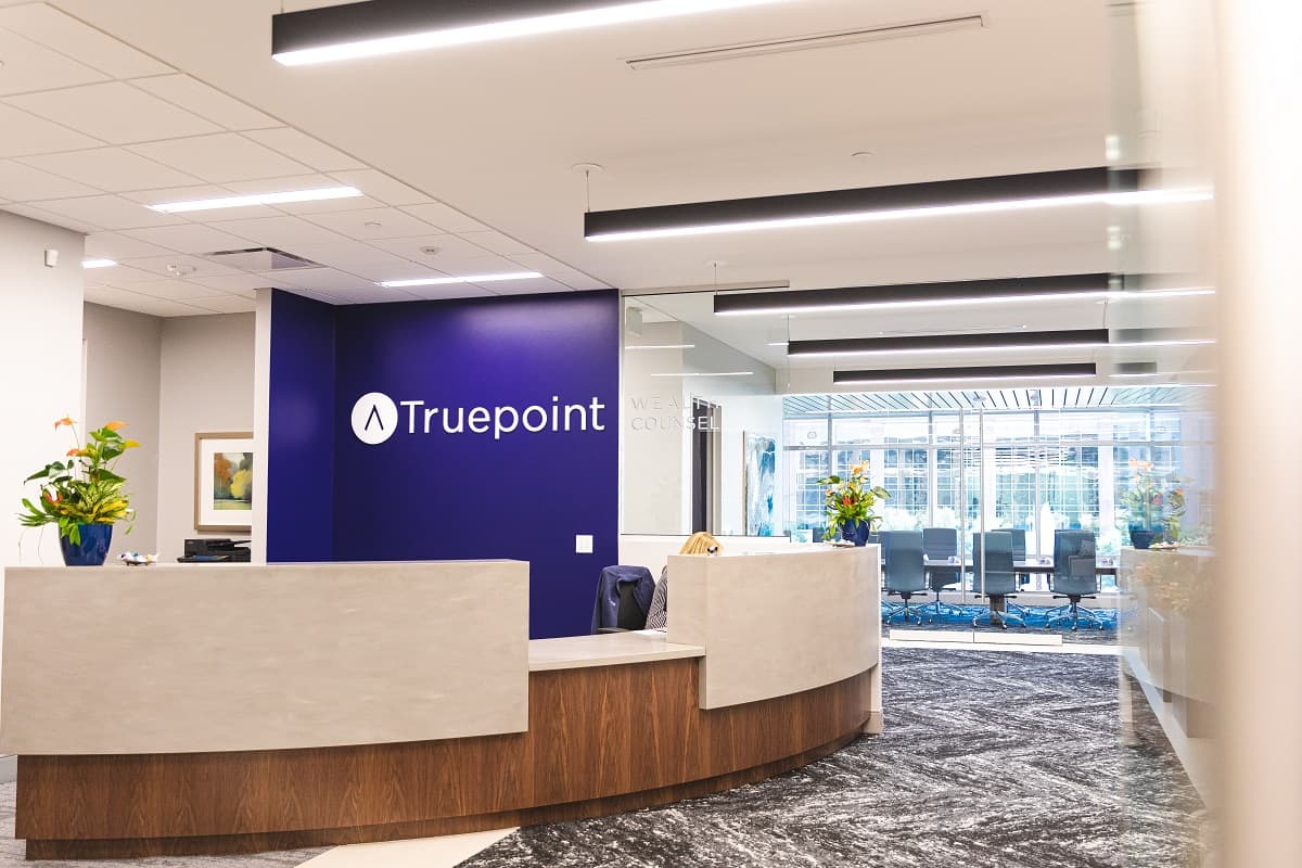 Modern office reception: curved desk, beige/dark blue; Truepoint on wall. Bright lights, large windows to conference room, green plants.