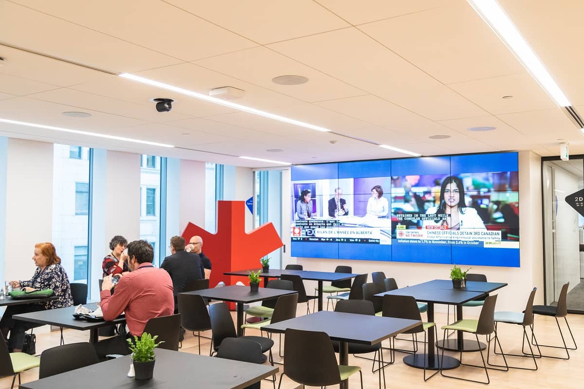 A modern cafeteria with people at tables, a news screen, white walls, large windows, black tables with plants, and a red sculpture.